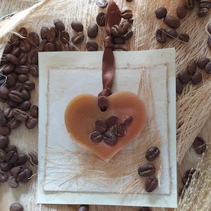 Könnte beinhalten: Ein herzförmiges, bernsteinfarbenes Seifenstück mit eingebetteten Kaffeebohnen auf einem strukturierten Papierhintergrund. Ein braunes Band ist an der Seife befestigt. Verstreute Kaffeebohnen umgeben die Seife und erzeugen eine rustikale, kaffeebezogene Ästhetik.