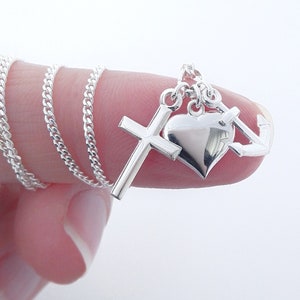 May include: A silver necklace featuring a cross, heart, and anchor pendant. The chain is a delicate curb link style. The pendant is highly polished and reflects light. The necklace is displayed against a white background.