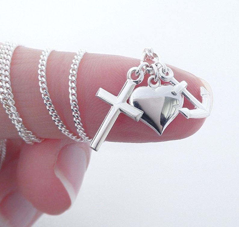 May include: A silver necklace featuring a cross, heart, and anchor pendant. The chain is a delicate curb link style. The pendant is highly polished and reflects light. The necklace is displayed against a white background.