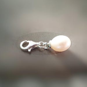 Puede incluir: Un charm con cierre de mosquetón plateado con una pequeña perla en forma de lágrima, de color blanquecino. El charm está conectado a la perla con una pequeña cadena. El fondo es gris liso.