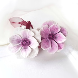 Puede incluir: Dos flores de tela blancas y moradas con una cinta burdeos. Las flores están hechas para parecer anémonas.
