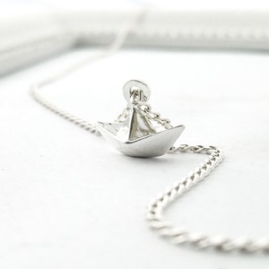May include: A silver necklace featuring a small, origami paper boat pendant. The chain is a delicate, twisted design.