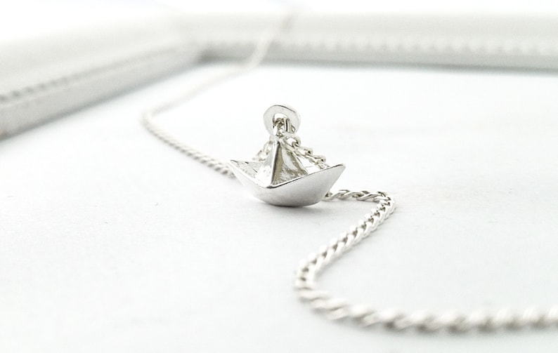 May include: A silver necklace featuring a small, origami paper boat pendant. The chain is a delicate, twisted design.