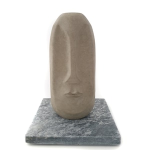 Puede incluir: Un jarrón de hormigón gris con forma de cara y un diseño minimalista. El jarrón está sobre una base de mármol gris.