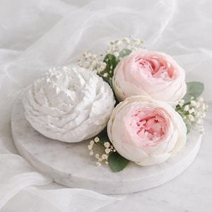 Op de afbeelding: Een marmeren serveerschaal met drie bloemen: één witte en twee roze. De roze bloemen hebben een lichtere roze kern. Groene bladeren en kleine witte bloemen omringen de bloemen. De schaal staat op een wit, getextureerd oppervlak.