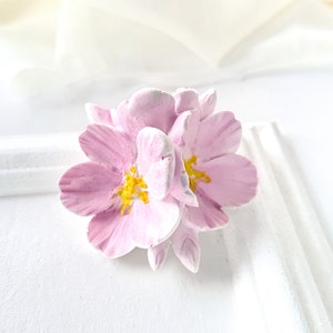 以下が含まれることがあります： 黄色い中心を持つピンクと白の3つの花が集まっている。花はポリマークレイで作られており、リアルな外観をしています。