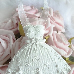 Könnte beinhalten: Weißer Porzellan-Brautkleid-Anhänger mit einem Satinband zum Aufhängen. Das Kleid hat ein Spitzenmuster und eine kleine Rose auf dem Oberteil.