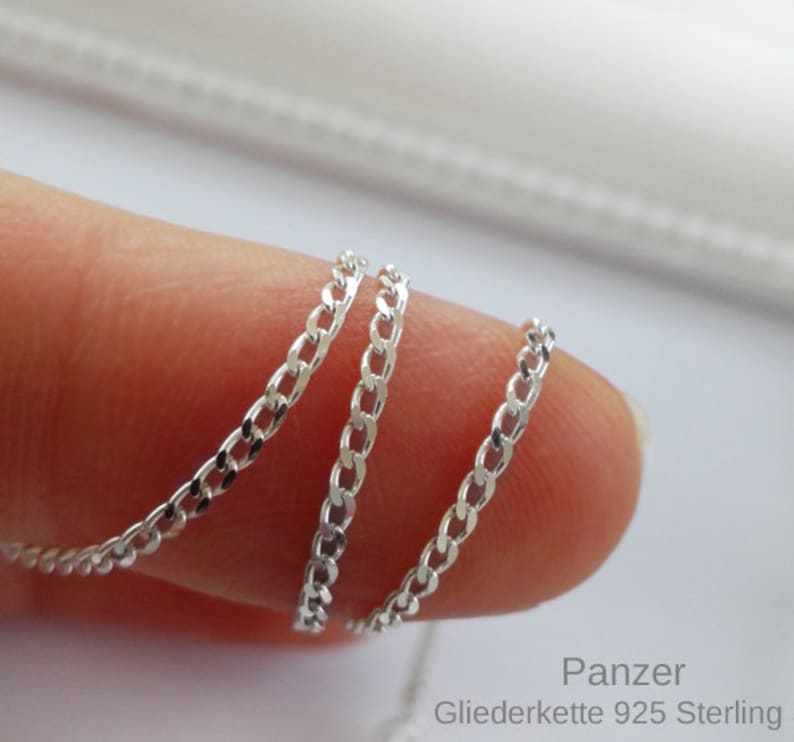 May include: A silver chain necklace with a curb link style. The chain is shown coiled on a finger. The text "Panzer Gliederkette 925 Sterling" is visible in the image.