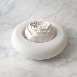 Pode incluir: Um difusor de botão de rosa de cerâmica branca fica em um anel de cerâmica branca. O difusor é projetado para conter óleos essenciais.