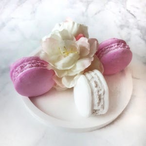 Könnte beinhalten: Ein weißes Keramiktablett mit drei Macarons. Zwei Macarons sind rosa und eines ist weiß. Eine weiße Blume mit rosa Akzenten liegt auf den Macarons.