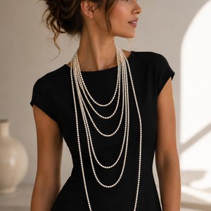 Puede incluir: Un collar de múltiples hilos que presenta varias capas de pequeñas perlas blancas redondas. El collar consta de varios hilos conectados de diferentes longitudes que caen sobre el pecho, usado sobre un vestido negro sencillo de manga corta.