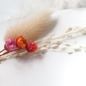Op de afbeelding: Een boeket gedroogde bloemen met een roze, oranje en rode bloem en wit pampasgras.