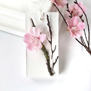 Puede incluir: Un adorno colgante de cerámica blanca con una flor rosa y una rama con musgo. El adorno tiene una cinta blanca.