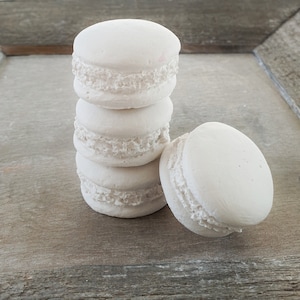 Può includere: Una pila di quattro macarons bianchi su una superficie di legno. I macarons sono disposti a forma di piramide, con il macaron più grande in basso e il macaron più piccolo in alto.