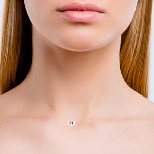 Può includere: Una delicata collana con un piccolo ciondolo argentato con la lettera "M". Il ciondolo è sospeso a una catena sottile e quasi invisibile. La collana è indossata intorno al collo.