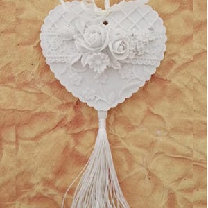 Puede incluir: Adorno de cerámica blanca en forma de corazón con un diseño de encaje y un motivo floral. El adorno tiene una borla blanca que cuelga de la parte inferior.