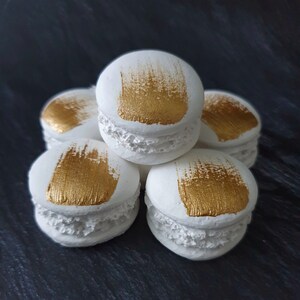 Könnte beinhalten: Fünf weiße Macarons mit goldenen Pinselstrichen auf der Oberseite. Die Macarons sind pyramidenförmig gestapelt.