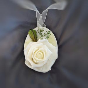 Könnte beinhalten: Ovaler, cremefarbener Blumenschmuck mit einer weißen Rose, grünen Akzenten und kleinen weißen Blüten. Ein weißes Band ist zum Aufhängen befestigt. Der Hintergrund ist ein dunkelgrauer Stoff.