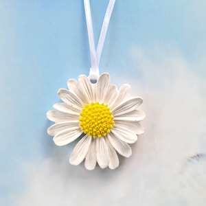 Puede incluir: Un amuleto de margarita blanca con un centro amarillo cuelga de una cinta blanca. El amuleto es un diseño simple y elegante.