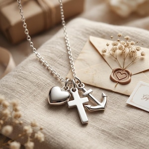 May include: A silver necklace with three charms: a heart, a cross, and an anchor. The necklace is displayed on a beige fabric, with a blurred background including a gift box and an envelope with a heart-shaped wax seal.