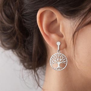 Puede incluir: Un pendiente colgante de tono plateado con un colgante circular que presenta un diseño recortado del árbol de la vida. El pendiente tiene un pequeño poste plano y redondo conectado al motivo circular mediante una corta barra metálica vertical.
