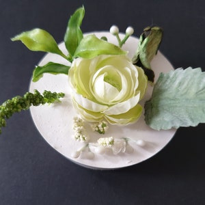 Puede incluir: Una flor blanca con hojas verdes y bayas blancas dispuestas sobre una base circular blanca.
