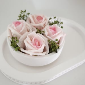 Puede incluir: Un cuenco de cerámica blanco lleno de cinco rosas de color rosa claro con centros de color rosa más oscuro y ramitas verdes. El cuenco está sobre una fuente ovalada blanca.