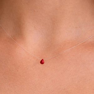 Pode incluir: Um colar com um único e pequeno pingente vermelho brilhante em forma de lágrima, suspenso por um fio de nylon fino e transparente que repousa sobre a pele, criando um efeito flutuante para o vibrante pingente de gota vermelha.