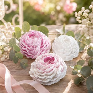 Könnte beinhalten: Drei seifenförmige Pfingstrosen in Rosa und Weiß liegen auf einer Holzoberfläche, umgeben von Grün und einem rosa Band. Die Seifen haben detaillierte Blütenblattstrukturen, die ein florales Thema hervorrufen. Ein Glas und eine Vase mit Blumen sind im Hintergrund.