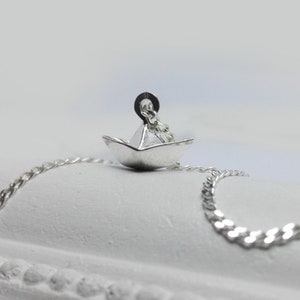 May include: A silver necklace with a small paper boat pendant. The chain is a curb chain style.