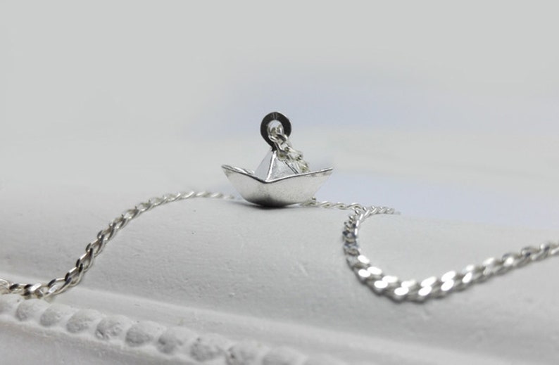 May include: A silver necklace with a small paper boat pendant. The chain is a curb chain style.