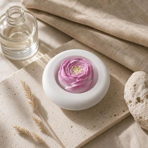 Puede incluir: Un plato de cerámica blanco, circular y de superficie lisa que contiene un objeto único en forma de flor rosa y blanca con un centro texturizado. El plato está colocado cerca de una botella de vidrio transparente y una piedra clara porosa.