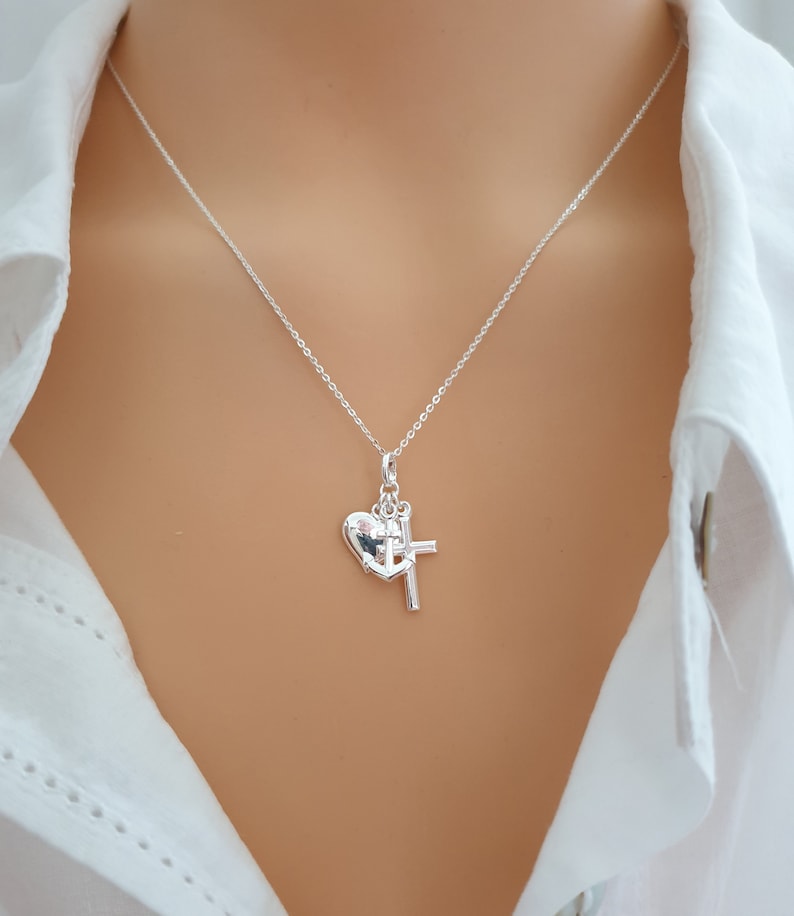 May include: A silver necklace featuring a heart-shaped pendant with a cross charm. The pendant is suspended from a delicate chain. The necklace is displayed against a white shirt.