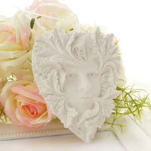 Könnte beinhalten: Weißer Keramik-Anhänger in Herzform mit einem detailliert gearbeiteten Gesicht und einem Blattdesign. Der Anhänger ist von weißen und rosafarbenen Rosen umgeben.