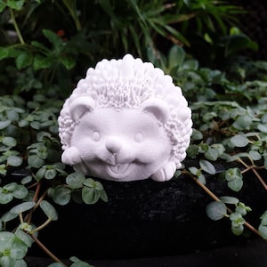 Puede incluir: Figurita de erizo de cerámica blanca con una cara sonriente. La figurita está sentada en un lecho de hojas verdes.