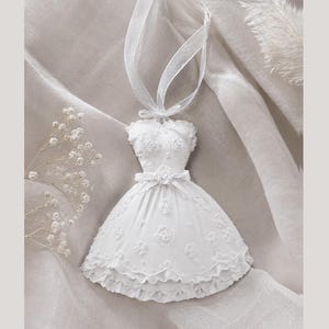 Könnte beinhalten: Ein weißes Hängeornament in Form eines trägerlosen Kleides mit herzförmigem Ausschnitt, einer taillierten Taille mit kleiner Schleife und einem ausgestellten Rock. Das Kleid ist mit geprägten Blumenmustern und einem Rüschensaum verziert.