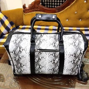 Peut inclure: Un sac de voyage rectangulaire avec un motif peau de serpent noir et blanc. Le sac présente des garnitures en similicuir noir, deux poignées supérieures avec des détails argentés et une poche zippée horizontale à l'avant. Base rigide à roulettes.