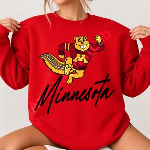 Op de afbeelding: Een felrood sweatshirt met ronde hals en een afbeelding van een cartoon gopher in een bordeauxrode en gouden trui met een groot M-logo. De gopher rent en het woord Minnesota staat eronder gedrukt in een vetgedrukt, zwart, cursief lettertype.