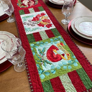 May include: A red, green, and white quilted table runner with a Christmas theme. The runner features three different designs: a wreath with a bird, a floral arrangement, and a cardinal with the word "Peace".