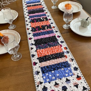 May include: A white table runner with a black and white border featuring a repeating pattern of stars and witch hats. The runner is made of fabric with a patchwork design of various Halloween-themed prints in orange, blue, pink, and black.