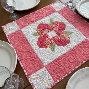 May include: A handmade quilted table runner featuring a central floral design in shades of red and green on a white background. The runner is bordered by a pink quilted fabric and floral patterned corners. The runner is placed on a wooden table, with white plates and glasses.