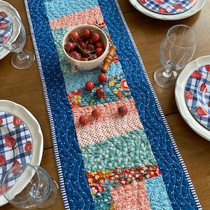 May include: A blue quilted table runner with white polka dots and a white border. The runner is decorated with colorful patchwork squares featuring floral patterns, red and white stripes, and a blue and white alphabet print. A bowl of red cherries sits on the runner.