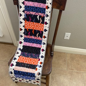 May include: A Halloween-themed table runner with a patchwork design featuring orange, pink, blue, and black fabric. The runner has a white border with a black and white pattern.