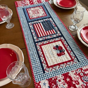 May include: A patriotic table runner with a patchwork design. It features a blue and white gingham pattern, red and white floral and star patterns, and squares with an American flag and floral embroidery. The runner is on a wooden table with red plates and glasses.