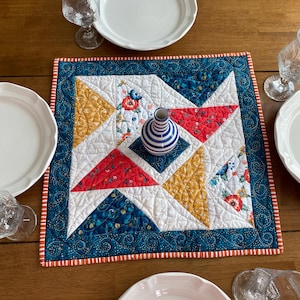 May include: A quilted table runner with a geometric pattern in red, yellow, and blue fabrics. The runner is placed on a wooden table with white plates and glasses.