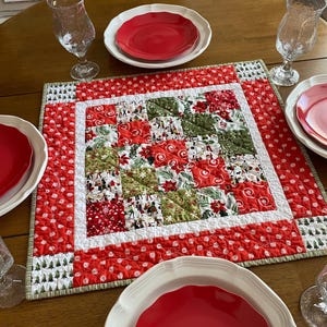 May include: A festive Christmas table runner with a patchwork design. The center features squares of red, green, and white fabric with holiday patterns. The border is red with white polka dots. Plates and glasses are visible.