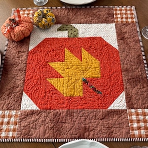 May include: A square quilted table mat featuring a central orange pumpkin block with a yellow maple leaf appliqué, surrounded by brown and white checkered borders. Two fabric pumpkins sit on the mat, one orange and one yellow floral.
