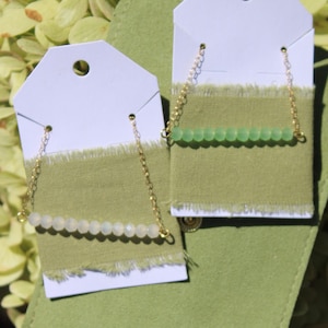 Puede incluir: Dos collares presentados sobre cartulina blanca con tiras de tela verde oliva deshilachadas. Un collar tiene una fila de cuentas blancas translúcidas y el otro una fila de cuentas verde claro, ambos unidos a finas cadenas de metal dorado.