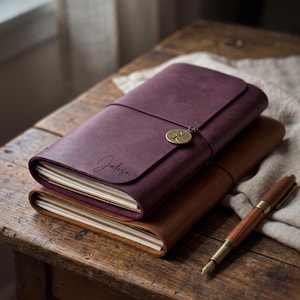 May include: A deep purple leather journal with the name Jalisa engraved on the cover, featuring a round brass tree charm attached to a thin cord. A second tan leather journal sits underneath, accompanied by a wooden fountain pen with brass accents.
