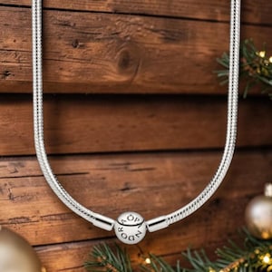 Puede incluir: Un collar de cadena de serpiente plateada con un cierre redondo grabado con la palabra PANDORA. La cadena tiene una textura suave y flexible con un acabado metálico pulido, formando una U sobre un fondo de madera oscura con agujas de pino.
